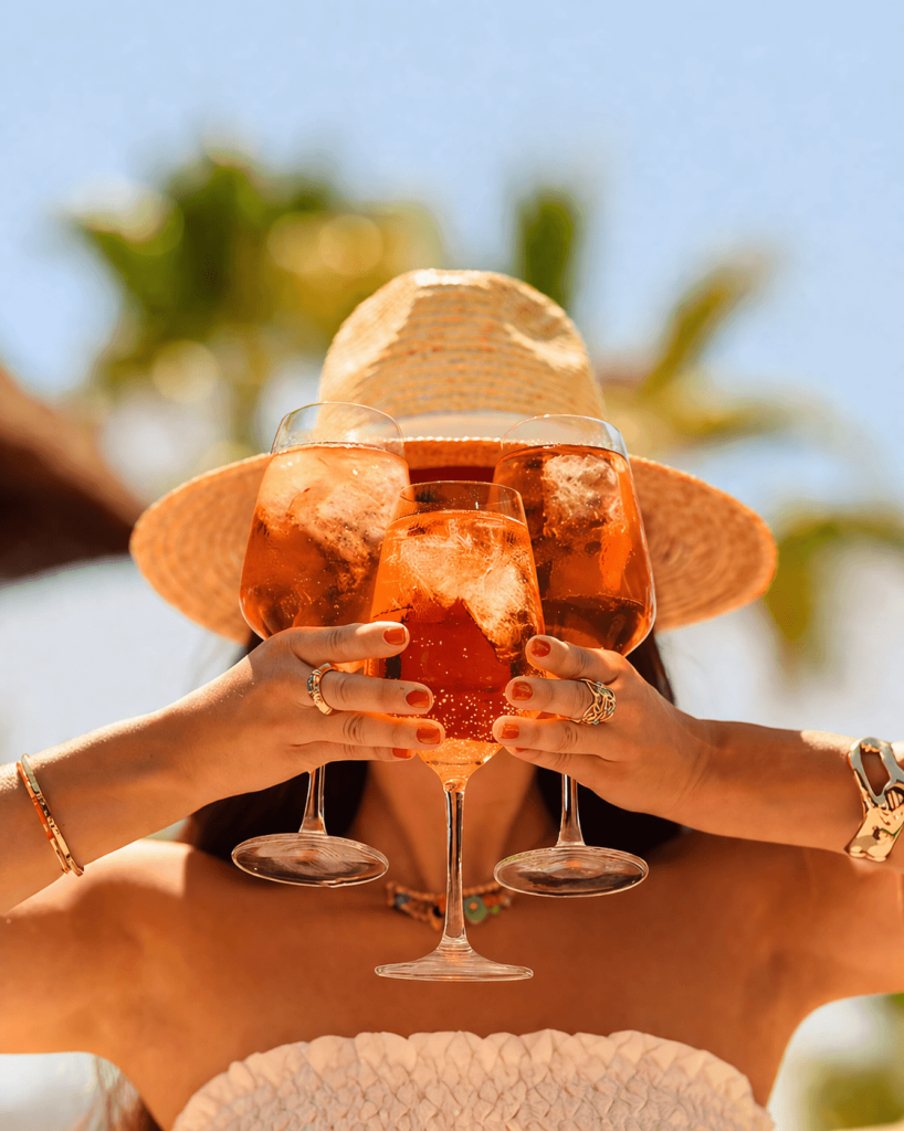 A person wearing a wide-brimmed straw hat playfully holds three large, vibrant orange Aperol Spritz cocktails in front of their face. The bright, sunlit background with blurred palm trees perfectly captures the refreshing "Splash of Aperol & Sunshine" tagline and the feel-good summer atmosphere of the Spritz Escape event