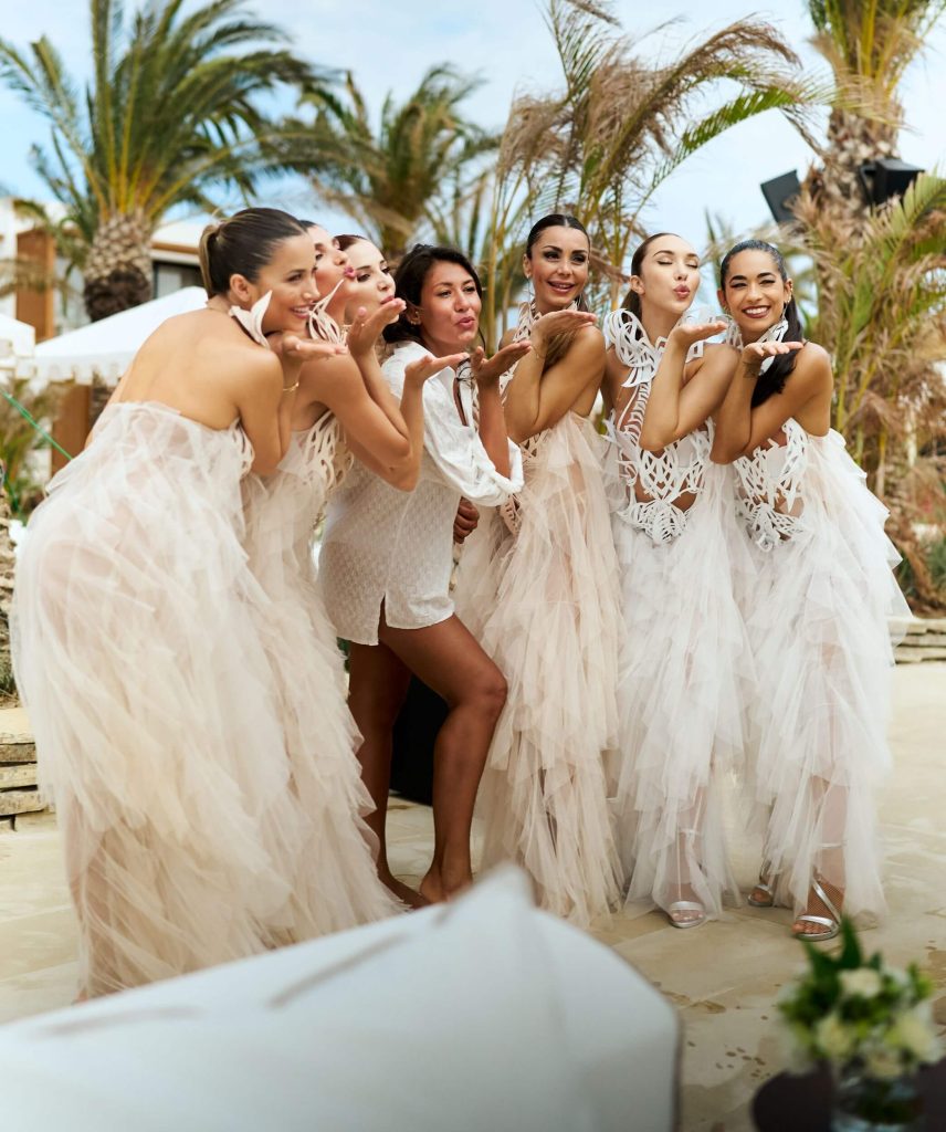 A lively group of women dressed in elegant white and cream attire smiling and blowing kisses to the camera, perfectly capturing the good energy and celebratory mood of the weekly Amazing Sundays event. The sunny outdoor setting with palm trees beautifully reflects the invitation to stay all afternoon and soak up the vibrant atmosphere