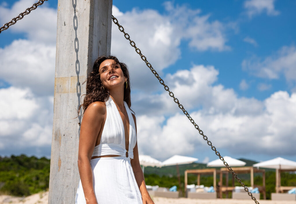 A women in a white dress leaning on an archway, on a sandy beach