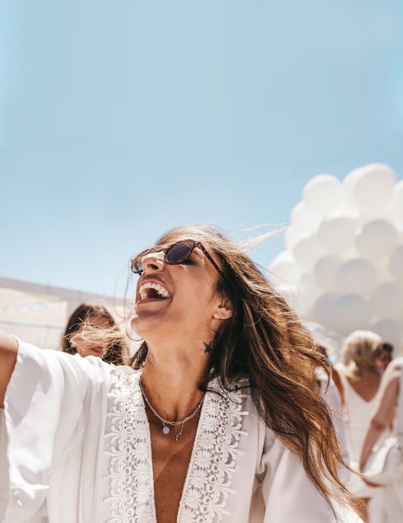 Smiling woman in white outfit enjoying a sunny outdoor celebration with white balloons in the background
