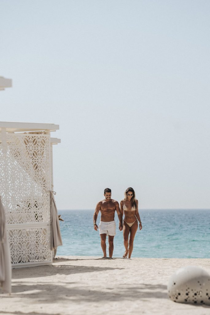 A smiling couple in swimwear walking hand-in-hand along a sandy beach, with a calm blue ocean backdrop and a white lattice cabana structure on the left
