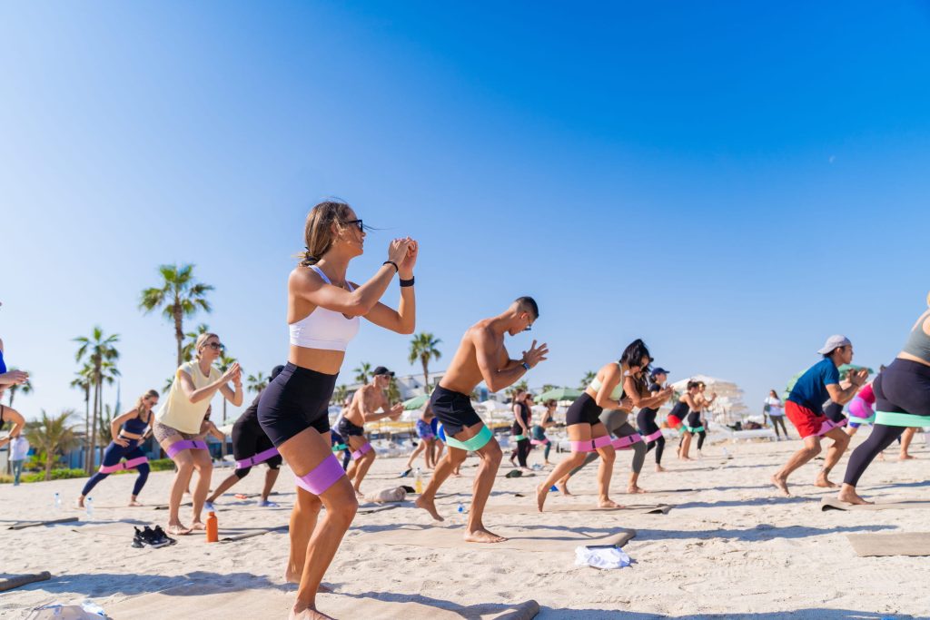 A group fitness class taking place on a sunny beach with palm trees in the background. Participants are performing squats on beige mats using colorful resistance bands around their thighs, with a woman in the foreground wearing a white sports bra and black shorts