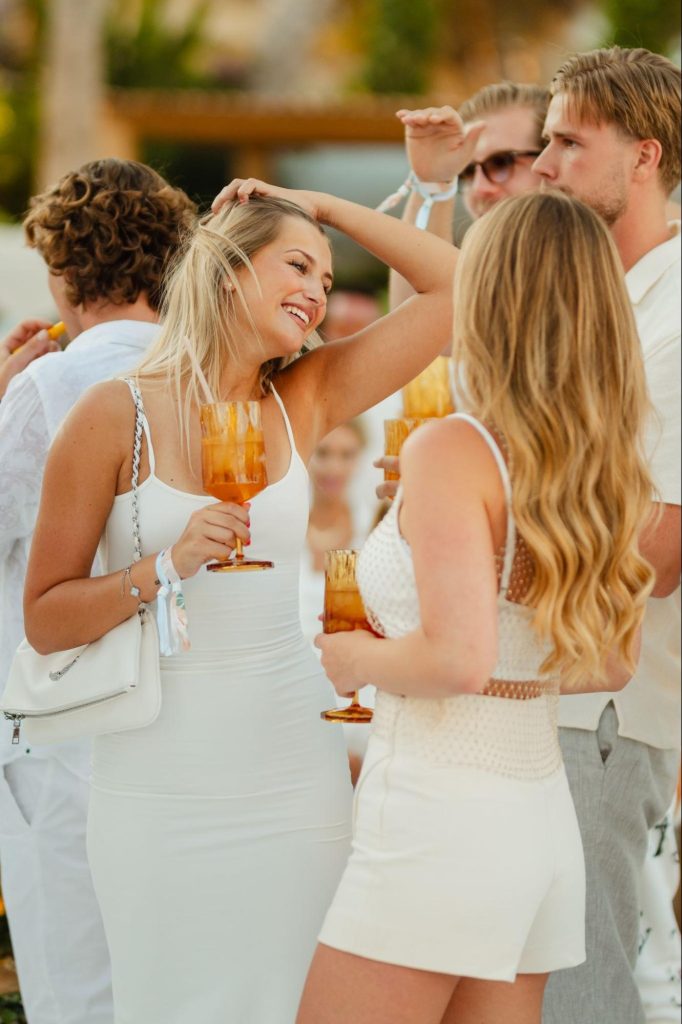 A group of guests enjoying a lively outdoor event, all dressed in chic white attire to match the legendary White Party theme. In the foreground, a smiling woman in a sleek white dress and a white crossbody bag chats joyfully with a friend while holding a vibrant orange cocktail, perfectly capturing the luminous and high-energy celebration of the evening