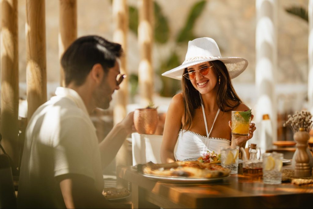 A smiling woman wearing a chic white wide-brimmed hat and stylish sunglasses holds a vibrant cocktail while dining outdoors with a companion. They are seated at a sunlit wooden table with food and drinks, perfectly capturing the relaxed yet fashion-forward atmosphere of the Amazing Sundays Brunch