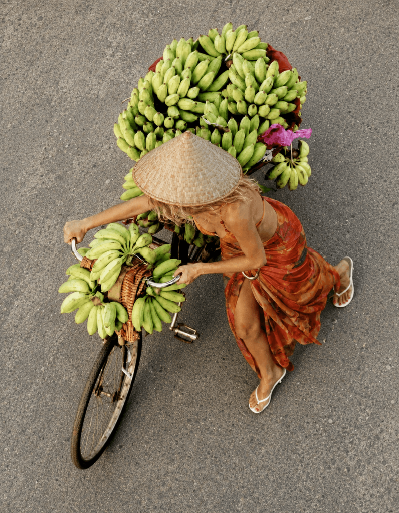 An overhead view of a woman wearing a flowing orange sarong and a traditional woven conical hat, pushing a bicycle heavily loaded with vibrant bunches of green bananas. This striking visual captures the colorful market energy and vibrant spirit of Thailand, perfectly setting the tone for the lively Bazaar by the Beach evening event at Nikki Beach Koh Samui.