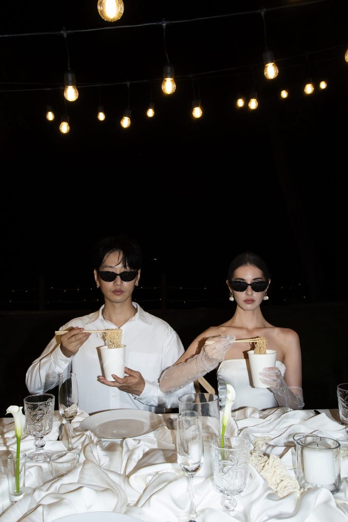 A promotional graphic for the Fatboy Izakaya pop-up at Nikki Beach Koh Samui, featuring a stylish couple dressed in formal white wedding-inspired attire and dark sunglasses. They are seated at an elegantly set table under night-time string lights, humorously eating noodles from branded "fatboy" cups with chopsticks. The banner overlay announces the event dates, July 31 to August 2, promising three days of bold bites, big beats, and unapologetic sizzle with Bangkok’s favorite modern izakaya