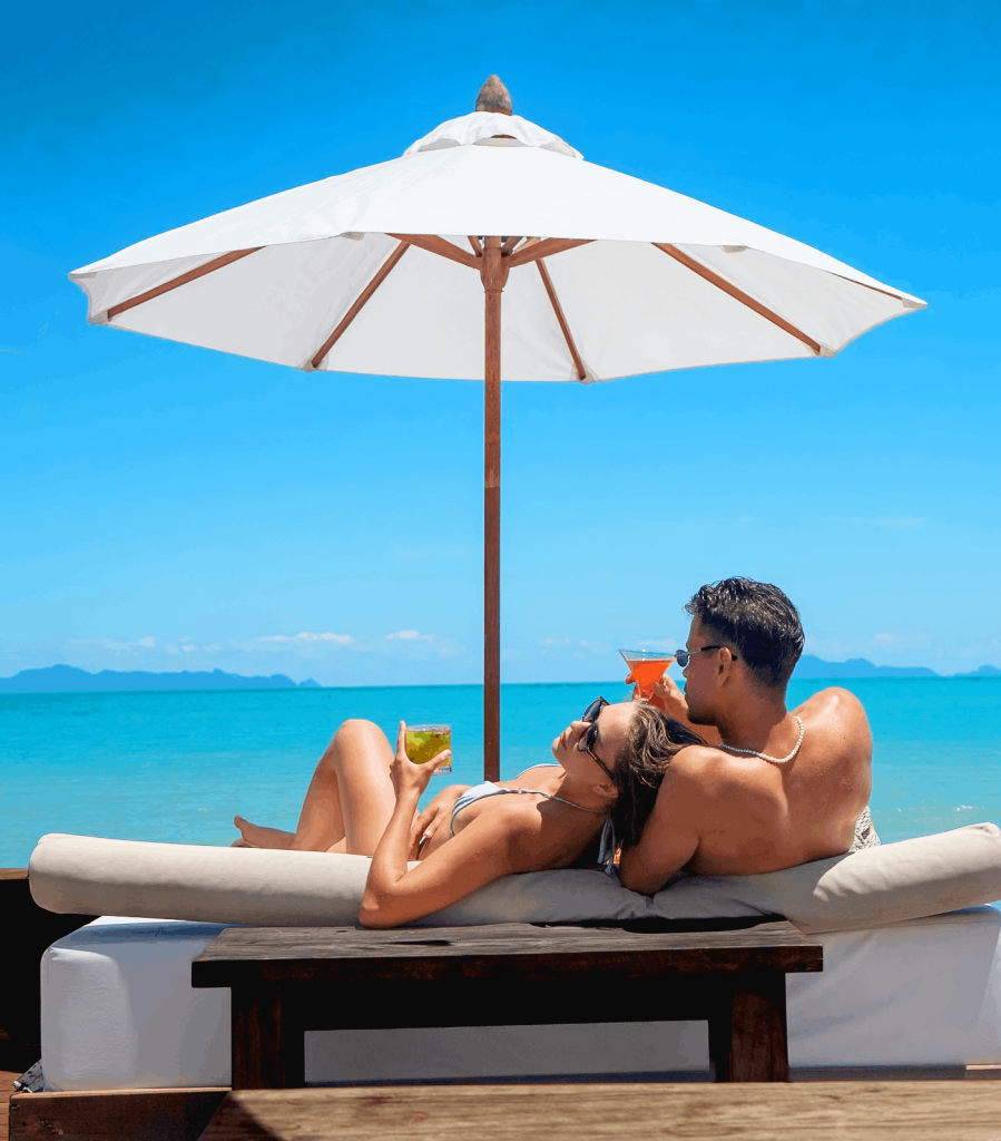 A couple relaxes on a plush white Sea View Sunbed under a white parasol, holding colorful cocktails against a stunning backdrop of bright turquoise water and a clear blue sky. This idyllic scene perfectly captures the effortless island vibes and tropical atmosphere awaiting guests who experience the Lancaster Sun Beauty collaboration at Nikki Beach Koh Samui