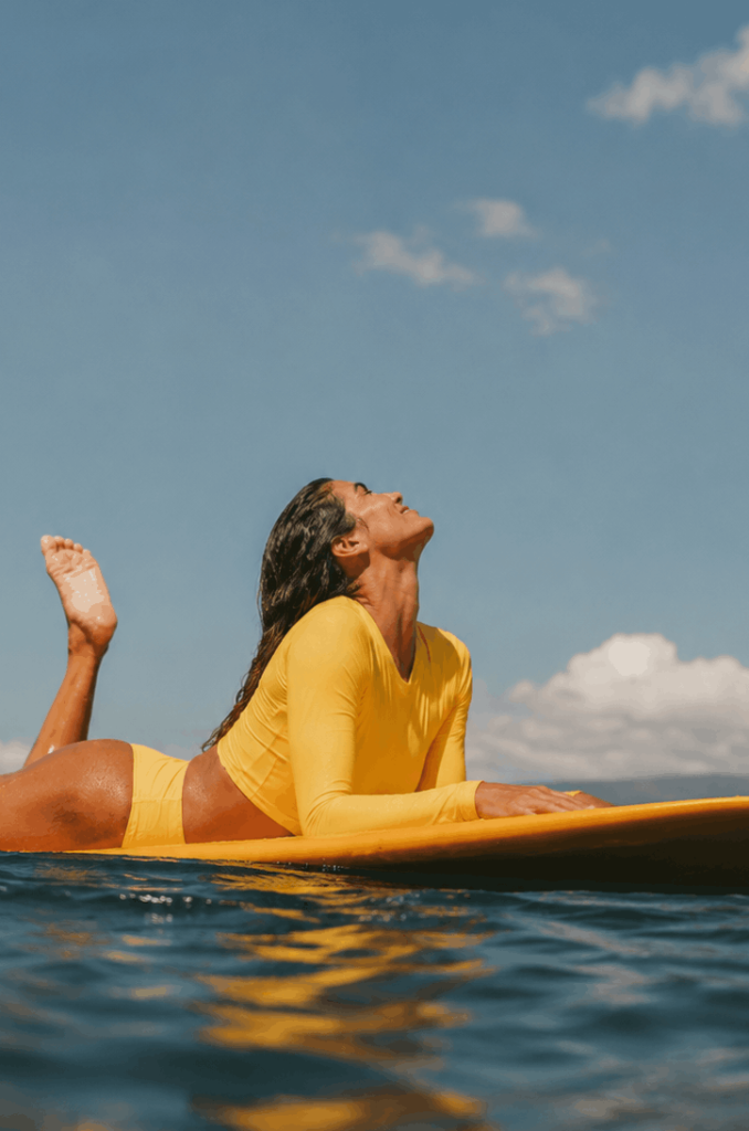 A woman wearing a bright yellow long-sleeve swimsuit relaxes on a yellow paddleboard in the ocean, tilting her head back to enjoy the warm sun against a clear blue sky. This serene moment on the water beautifully captures the restorative paddleboard yoga and rejuvenating island lifestyle experienced during the exclusive Wellness Reset with Roxy event at Nikki Beach Koh Samui.