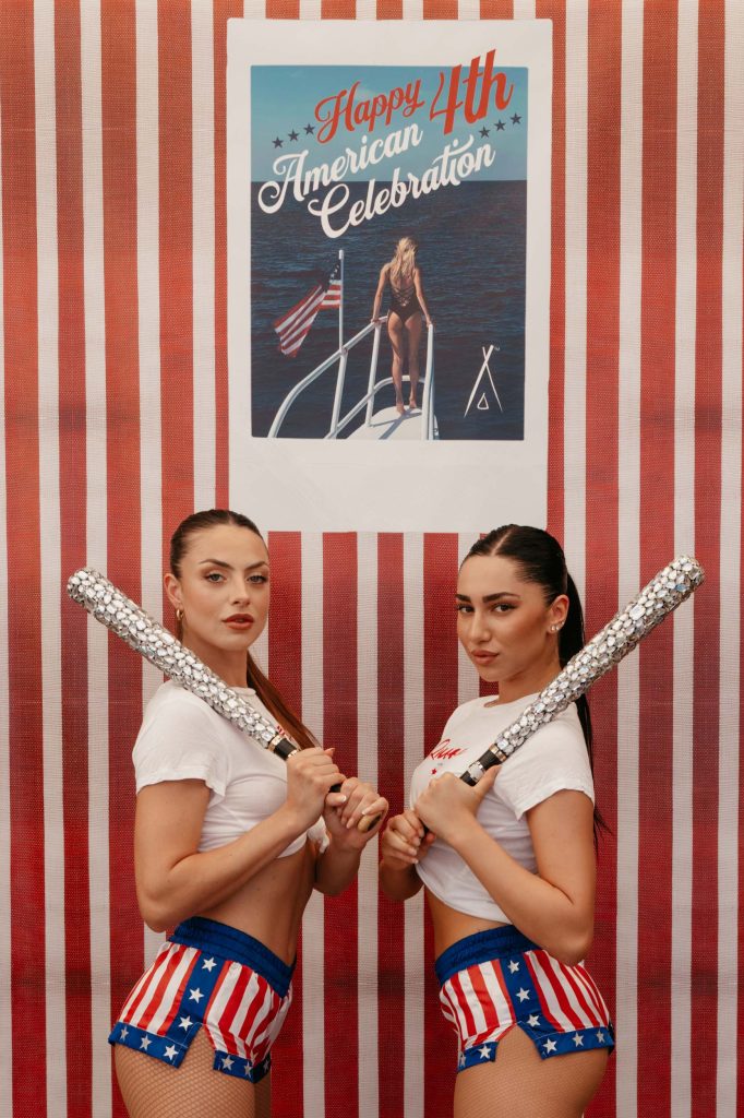 Two women posing in American flag clothing, holding sparkly baseball bats in front of a "Happy 4th American celebration" poster