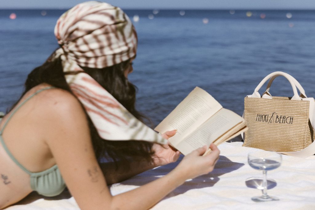 A lady reading a book laying on a blanket, next to a Nikki Beach bag