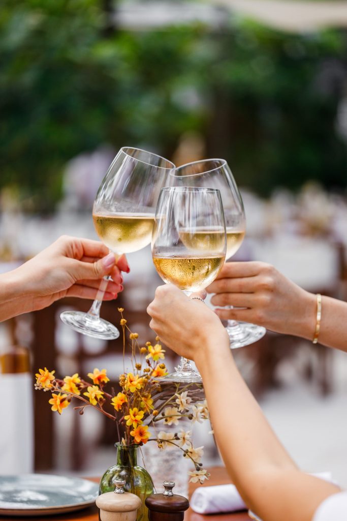 Three hands raise and clink glasses of pale rosé wine over a dining table decorated with small yellow and orange flowers. This elegant, sunlit toast perfectly captures the vibrant beachfront dining atmosphere of the National Rosé Day celebration at Nikki Beach Miami Beach