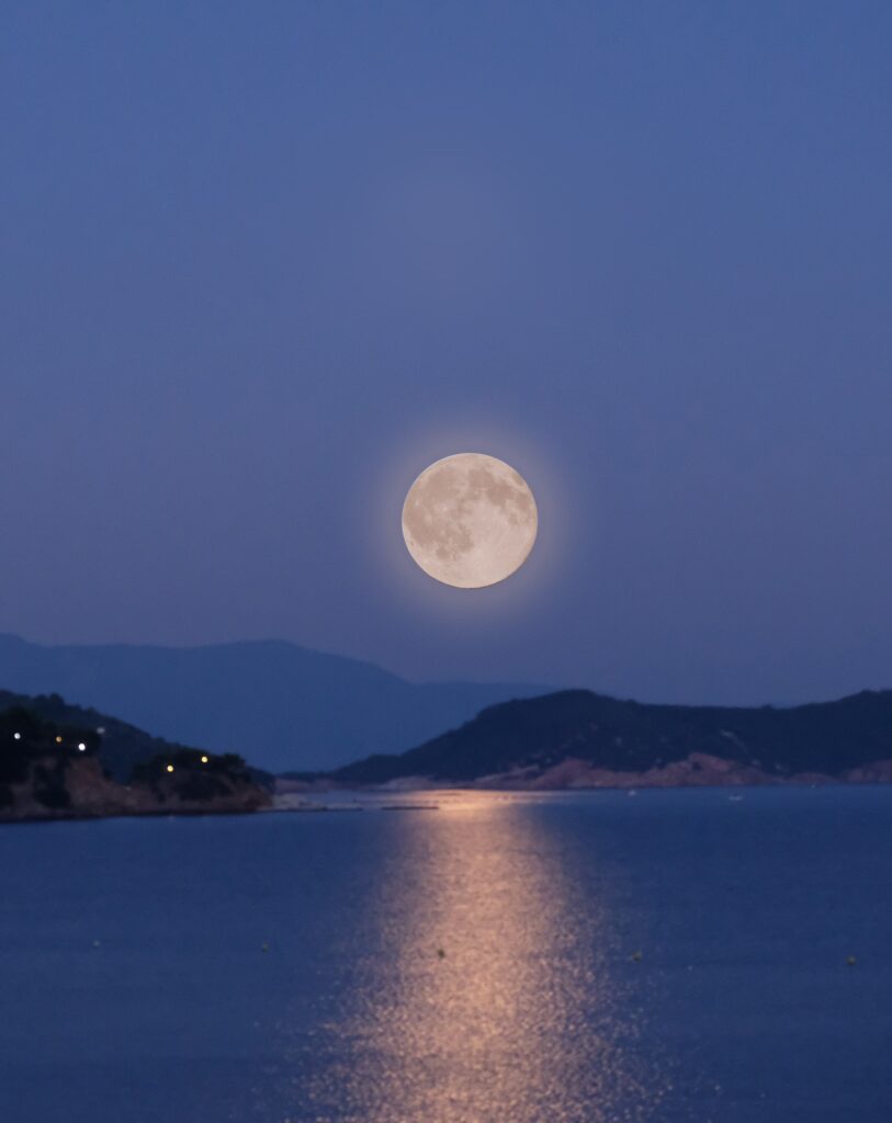 A luminous full moon casts its glow over the sea, framed by majestic mountains, setting the stage for a magical evening