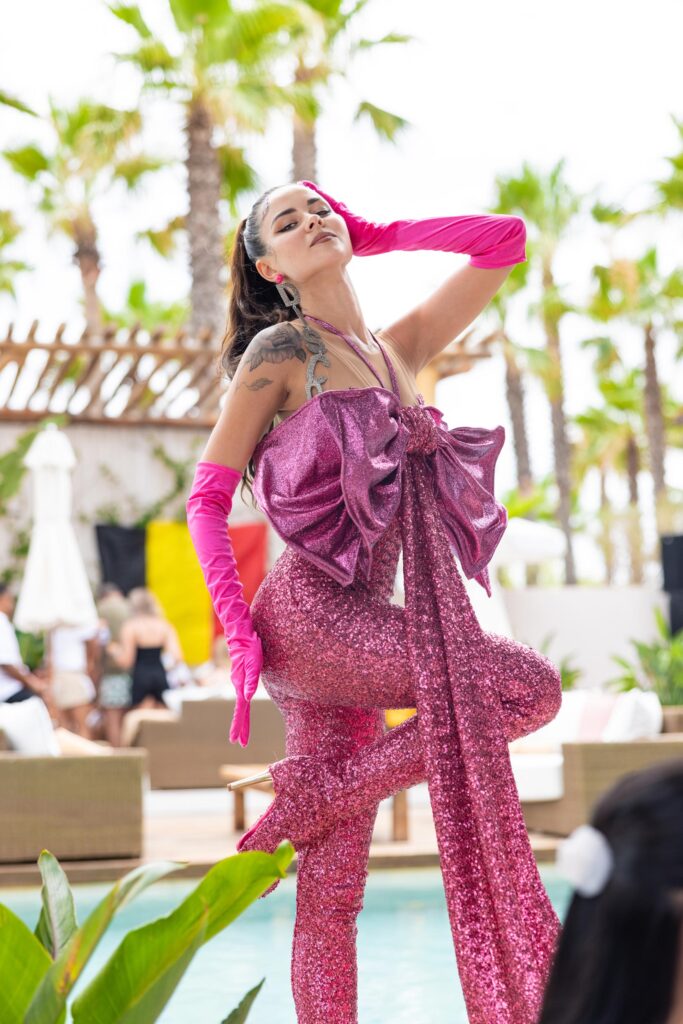 A lady posing with leg up and hand on head, wearing a sparkly pink jumpsuit