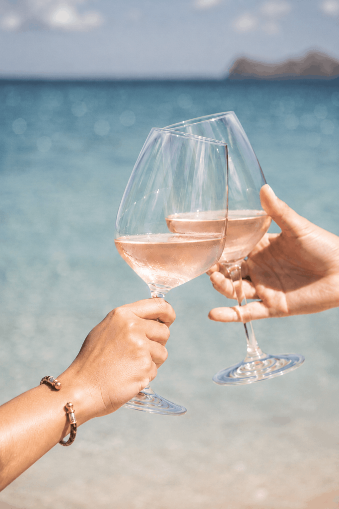 Two hands clink elegant wine glasses filled with chilled pale rosé against the stunning, sparkling backdrop of the turquoise Aegean Sea. This refreshing seaside toast perfectly captures the relaxing, sun-soaked atmosphere of the exclusive Rosé Escape poolside experience in partnership with Costa di Rosé